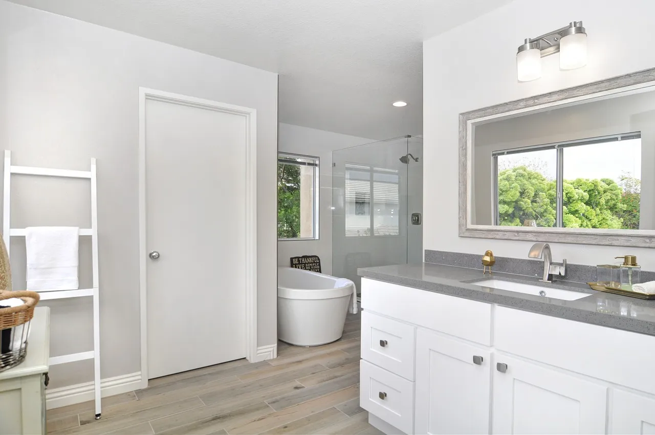 A modern bathroom features a white vanity with a gray countertop, a mirror above, and a sink. To the side, there's a standalone bathtub next to a window, and a door leads to a shower area. A ladder towel rack holds folded towels. Light wood flooring completes the look.