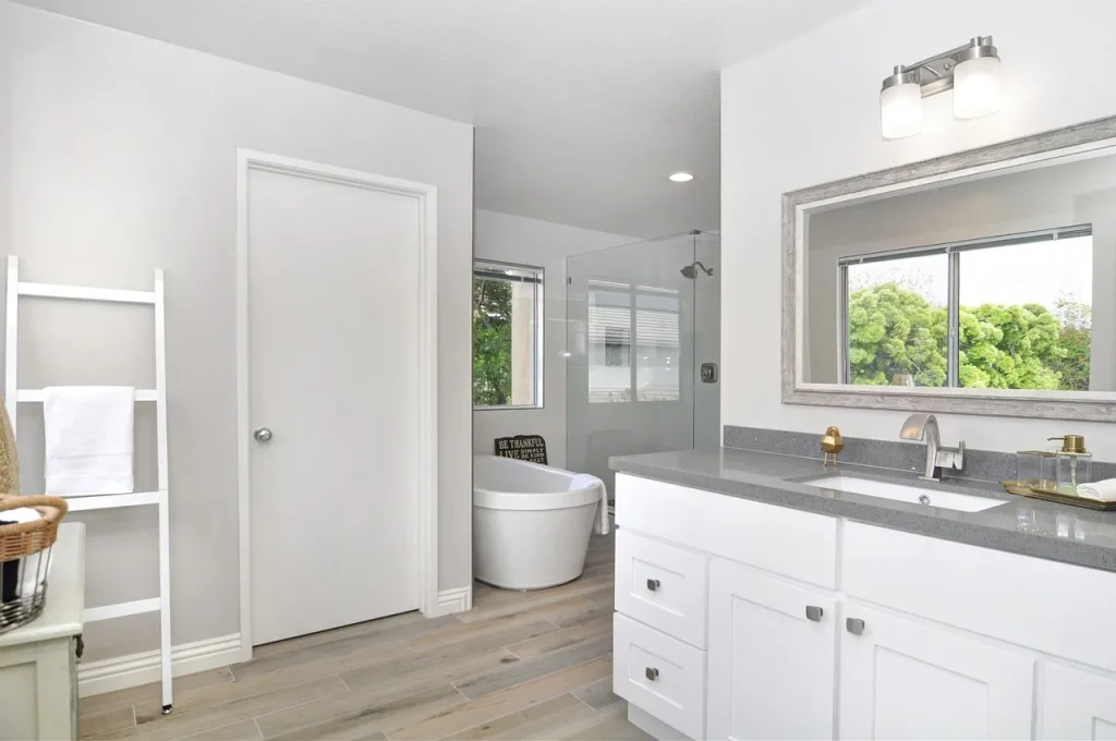 Modern bathroom with a white vanity, gray countertop, and large mirror. A white standalone bathtub is near a window with green foliage outside. A ladder shelf with towels and a wicker basket are on the left. Light wood flooring adds warmth.