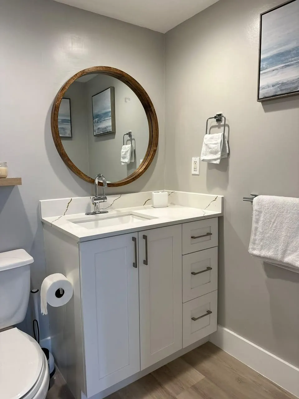 A modern bathroom features a round mirror above a white vanity with a marble countertop. The vanity has three drawers and a toilet nearby. Towels hang on the wall, and a framed abstract picture adds decor. The walls are light gray.