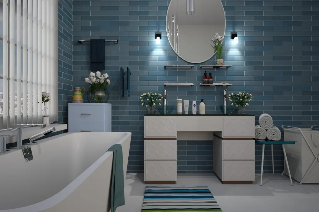 Modern bathroom with a freestanding bathtub, blue tiled walls, and a circular mirror above a vanity. Shelves hold plants and toiletries. A striped rug lies on the floor, and a rack with towels is on the right. Natural light filters through blinds.