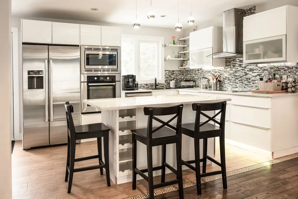 A modern kitchen with white cabinets, a glossy island, and black bar stools. Stainless steel appliances, including a double oven and refrigerator, are built into the cabinetry. The backsplash features a textured gray tile pattern.