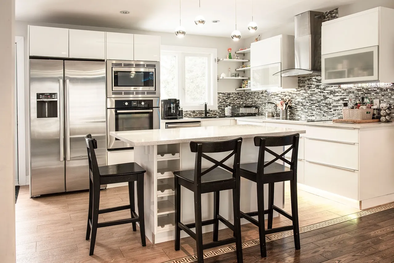 A modern kitchen with white cabinets, a glossy island, and black bar stools. Stainless steel appliances, including a double oven and refrigerator, are built into the cabinetry. The backsplash features a textured gray tile pattern.