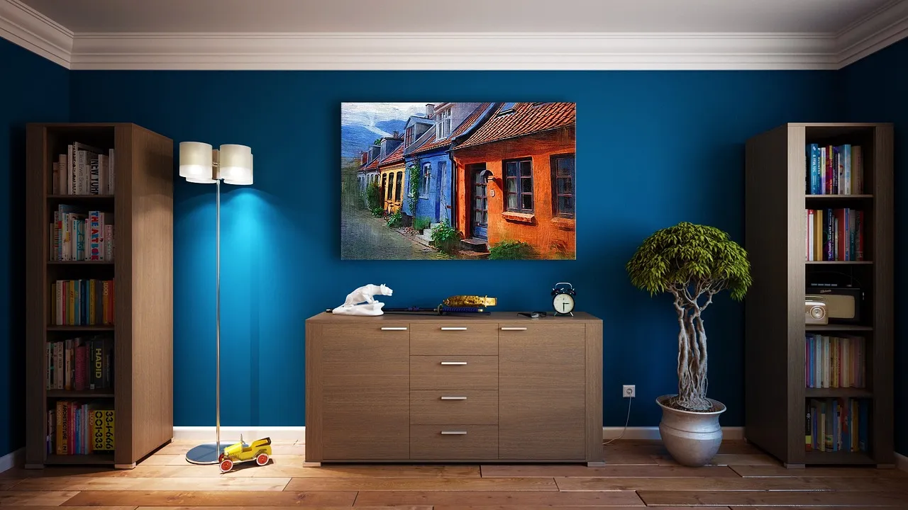 A cozy room with blue walls features a wooden dresser with drawers, a potted plant, and a tall lamp. A colorful street painting hangs above the dresser, with bookshelves on either side. A few toys are scattered on the floor.