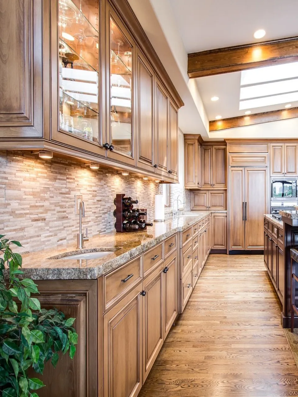 A spacious kitchen with wooden cabinets, granite countertops, and a stone backsplash. Skylights illuminate the room, and a wine rack is placed near the sink. A potted plant adds greenery to the hardwood floor setting.