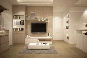 Modern living room with wooden accents, featuring a wall-mounted TV, bookshelves with decor items, a white coffee table with vases on a gray rug, and recessed lighting. The design is minimalistic with neutral tones and sleek furniture.