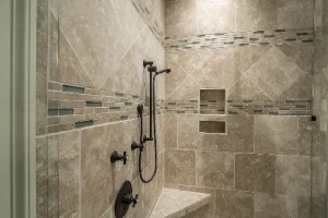 A modern shower with beige stone tiles, featuring geometric patterns. There is a built-in bench and a wall niche. The showerhead and controls are dark metal, and accents include blue and green mosaic tile strips.