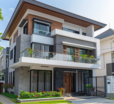 Modern three-story house with a mix of wood, stone, and glass exteriors. It features large windows, a second-floor balcony with plants, and a well-maintained lawn with shrubs. The entrance has a wooden door and sleek design elements.