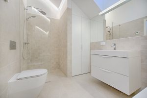 Modern bathroom with neutral tones featuring a wall-mounted toilet, a walk-in shower with glass enclosure, and a white vanity with sink and storage drawers. Large mirror and bright overhead lighting enhance the spacious feel.