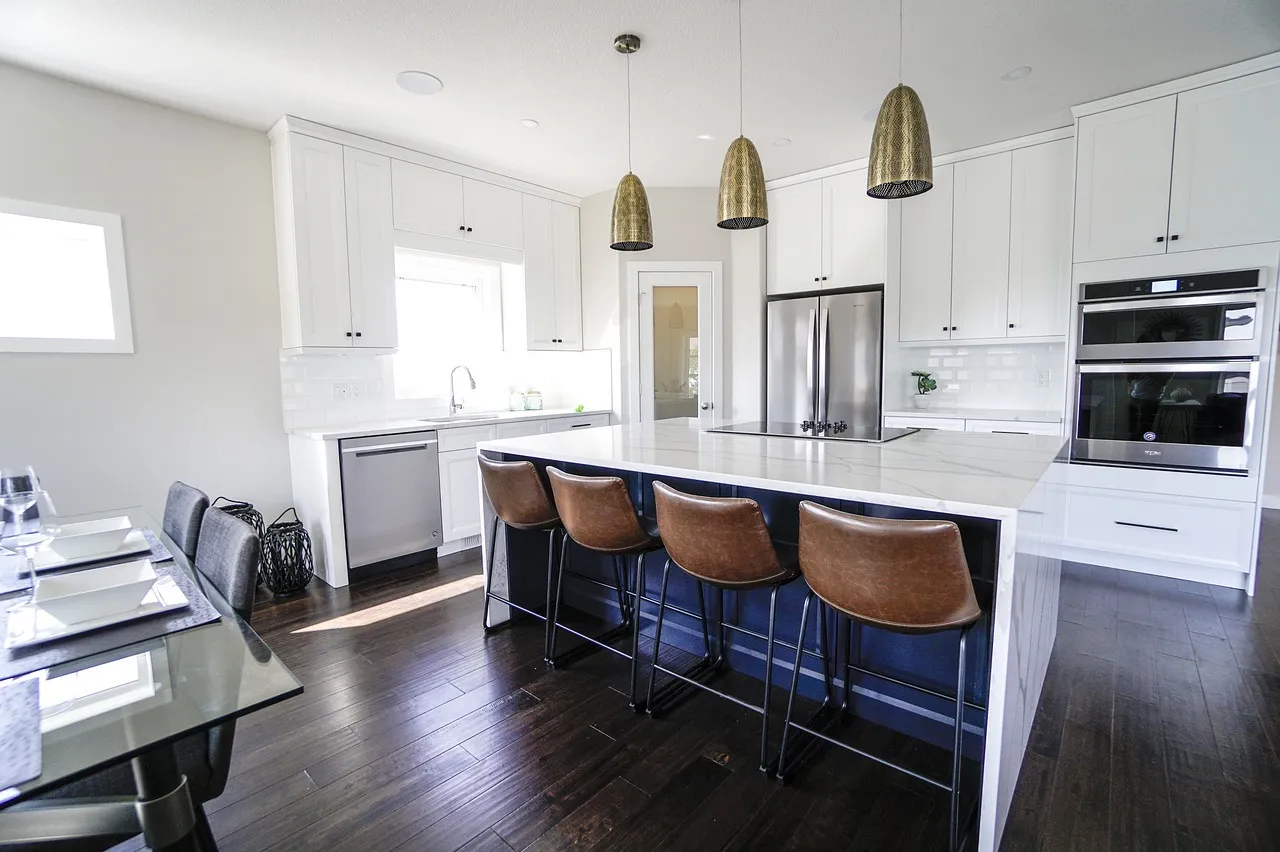 Modern kitchen with white cabinets and dark hardwood floors. A large island with a white countertop is surrounded by brown leather barstools. Stainless steel appliances, including a double oven and fridge, are integrated. Pendant lights hang overhead.