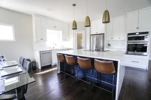 Modern kitchen with white cabinets, stainless steel appliances, and a dark blue island. Three brown leather bar stools line the island. Gold pendant lights hang above. A dining table with gray chairs is in the foreground on dark hardwood floors.