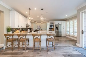 Spacious kitchen with a large island featuring four wooden chairs. White cabinets, stainless steel appliances, and pendant lights enhance the modern design. Light wood flooring and large windows provide a bright, airy atmosphere.