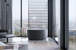 A modern bathroom with a round freestanding tub near tall windows. Striped blinds partially cover the view of rolling hills. A gray chair and a towel stand are nearby, with light wood flooring complementing the minimalistic design.