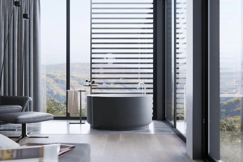 Modern bathroom featuring a sleek, round tub near large floor-to-ceiling windows with horizontal blinds. The room is bright, with soft lighting and a view of mountains outside. There's a plush chair and towel rack nearby, adding to the serene atmosphere.