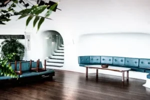 A modern, minimalist room with blue curved bench seating, a wooden coffee table, and an overturned chair. A staircase with a sleek design is visible, leading upward. A large potted plant adds greenery to the space, which has wooden flooring.