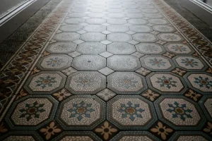 A corridor with intricate tile flooring showcases octagonal patterns in shades of blue, gray, and beige. The floral and geometric designs lend a vintage and artistic look to the dimly lit space.