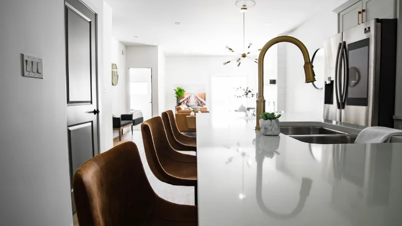 Modern kitchen with a white countertop island, brown bar stools, gold faucet, and stainless steel appliances. In the background, a living area with a tan sofa, plants, and wall decor is visible, creating a bright and inviting space.